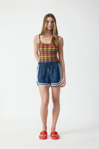 Full-length studio front view of a blonde model wearing mid-rise denim blue chambray shorts with cream binding along the dolphin hem, styled with a plaid camisole top and red slide sandals.