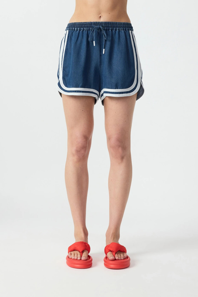 Studio front view from the waist down of relaxed denim blue chambray running-style shorts with an elastic drawstring waist and contrast cream hem piping.