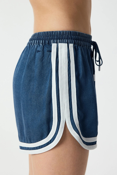 Close-up studio side view focusing on the layered cream piping binding, curved dolphin side hem, and elastic waistband on denim blue chambray shorts.