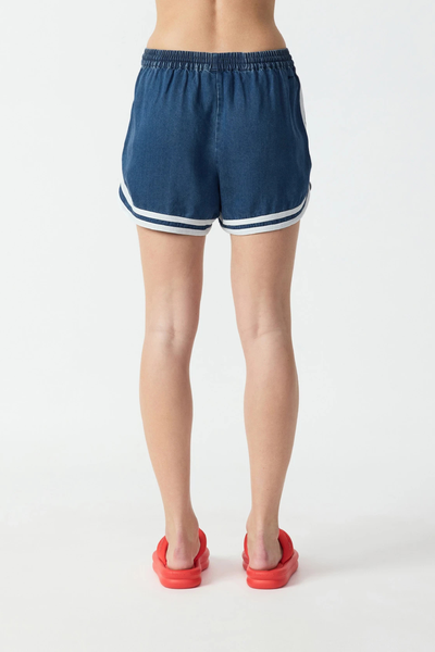 Studio rear view from the waist down of relaxed denim chambray shorts with a gathering back waistband, curved cream hem binding, and red sandals.