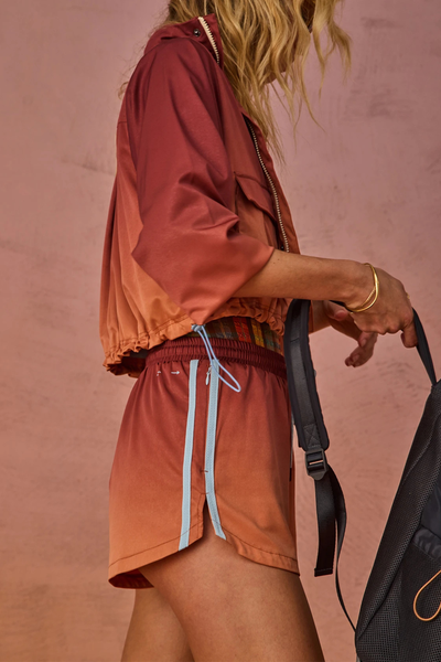 Outdoor side profile close-up of a model wearing sunset ombre red-to-orange run shorts with light blue side-seam mesh tape, paired with a matching jacket and carrying a backpack.