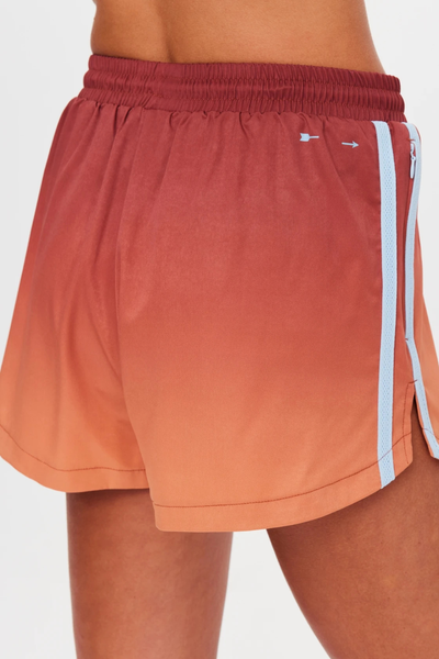 Close-up rear detail shot highlighting the printed white arrow logo, elasticated maroon waistband, and light blue side tape on red-to-orange ombre running shorts.