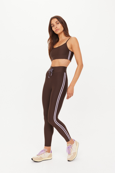 Full-length studio shot of a model wearing the high-performance Oxford 25-inch Midi Pant in cocoa brown. This side-on view highlights the supportive mid-rise waist, the double lilac binds on the side seams, and the midi crop length grazing just above the ankle.