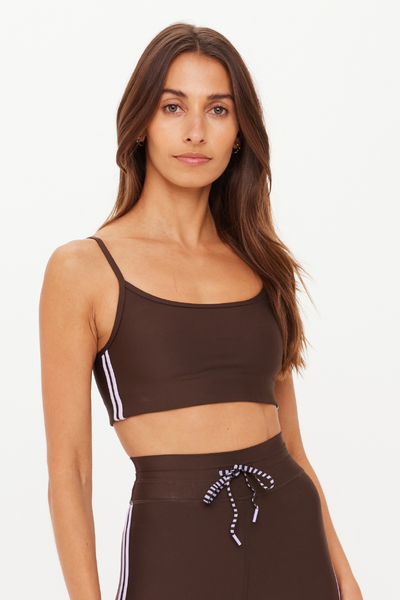 A close-up studio portrait of a model wearing the Oxford Hutton Scoop Bra in cocoa brown. The activewear crop top features a classic scoop neckline, thin adjustable shoulder straps, and subtle double lilac stripes detailing the side seam.