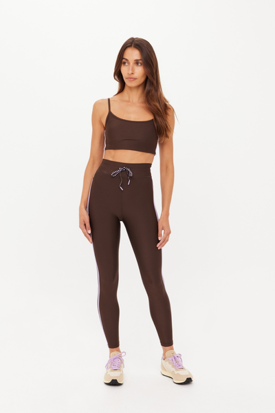Full-length studio shot of a model wearing the matching activewear set of the cocoa brown Oxford Hutton Scoop Bra and high-waisted Oxford leggings. The set is detailed with lilac side stripes, a striped drawstring waist, and is paired with light beige sneakers with lavender laces.