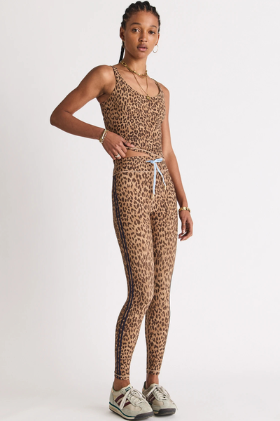 Studio three-quarter front view of a model wearing mid-rise 25-inch leopard print midi leggings with contrast navy side binding and a blue drawstring waist, paired with a matching leopard tank top and sneakers.