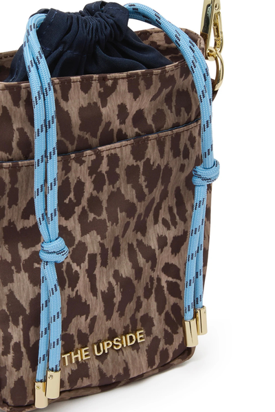 Close-up studio detail shot of a leopard print recycled polyester crossbody bag featuring a navy internal drawstring closure with light blue paracord ties, a front slip pocket, gold hardware, and a gold metal "THE UPSIDE" logo decal.