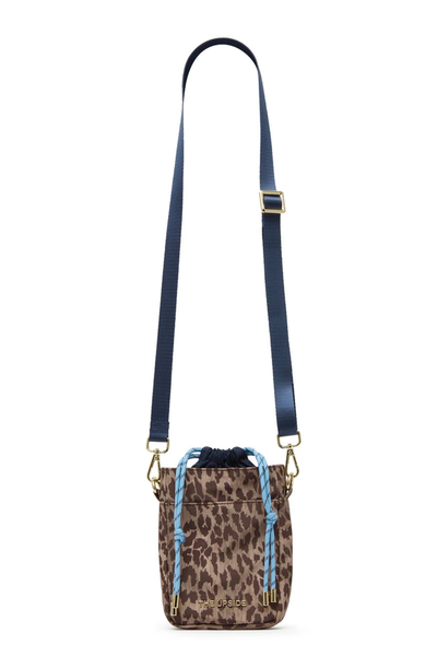 Studio product front view of a small leopard print crossbody bag made from recycled polyester, featuring light blue paracord drawstrings, a front slip pocket, gold "THE UPSIDE" metal logo decal, and an adjustable, removable navy webbed shoulder strap.