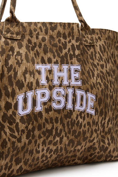 Close-up studio detail shot of the organic cotton leopard print fabric featuring a bold printed collegiate-style logo reading "THE UPSIDE" in lilac.