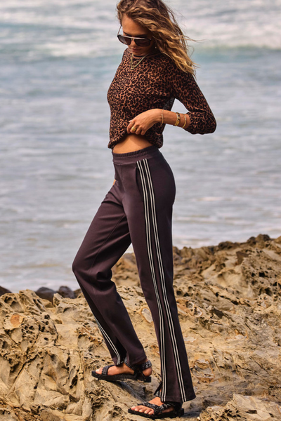 An outdoor campaign shot featuring a model crouching on coastal rocks, highlighting the flexibility and soft suiting texture of the liquorice grey Petra Flare Pant while showing the side-stripe panel detailing in profile.