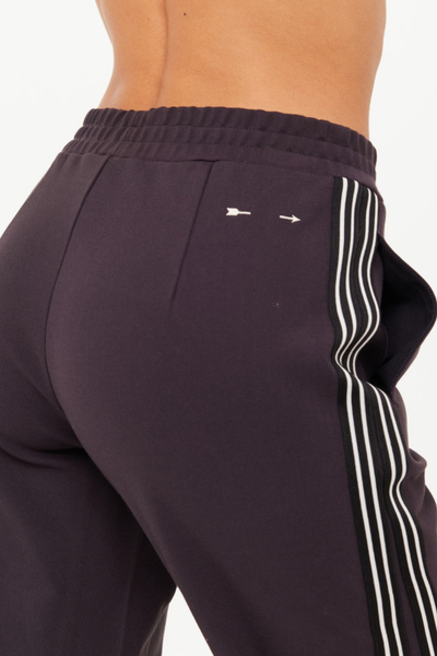 A macro close-up studio shot focusing on the back upper right hip of the liquorice grey track pants, detailing the gathered elastic waistband texture, a side pocket opening, and the signature cream embroidered arrow branding motif next to the black and cream side-stripe tape.
