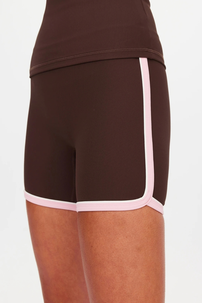 Close-up view of the side profile of the chocolate brown spin shorts, showcasing the curved hem and contrasting cream and pink side stripe binding.