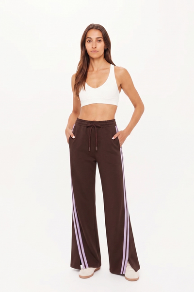 Full-length studio shot of a model wearing the high-waisted Roma Pant in cocoa brown, styled with a simple white sports bra and beige sneakers. The wide-leg track pants feature sporty lilac side stripes and front-split ankle hems.