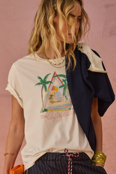 Half-length studio portrait of a blonde model with cuffed sleeves, wearing a cream organic cotton graphic t-shirt printed with a tropical palm tree illustration, styled with a navy sweater draped over her shoulder.