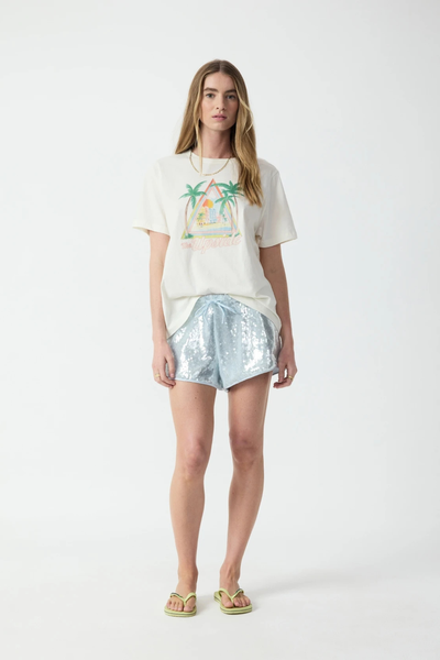 Full-length studio front view of a blonde model wearing a regular fit cream organic cotton graphic t-shirt, light blue sequin shorts, and lime green flip-flops.