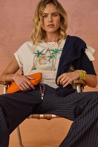 Outdoor seated lifestyle portrait of a blonde model wearing a cream organic cotton t-shirt with rolled sleeves, navy pinstriped drawstring pants, holding an orange pouch bag.