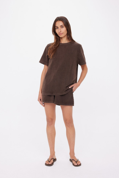 A three-quarter studio shot showing a model standing with one hand in her pocket, demonstrating the soft drape, comfortable shoulder construction, and washed textural finish of the deep cocoa brown cotton fabric.