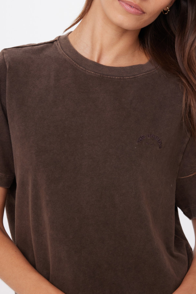 A detailed macro close-up studio shot of the upper chest area, highlighting the fine texture of the soft brushed recycled cotton fabrication, the clean stitched construction of the ribbed crew neck, and the arched "THE UPSIDE" tonal embroidered horseshoe branding motif.