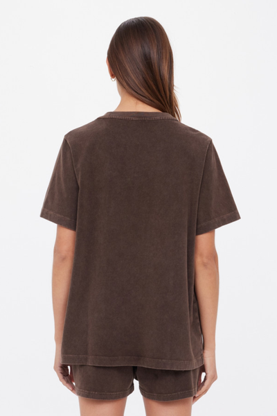 A studio product shot from the back capturing a model wearing an oversized, short-sleeve crewneck t-shirt in a washed cocoa dark brown shade. The image highlights the relaxed silhouette, standard crew neckline, clean shoulder seams, and the soft, textured appearance of the brushed cotton fabrication, styled as a matching set with lounge shorts against a plain white background.