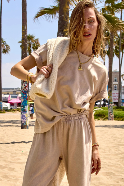 Lifestyle beach campaign photo of a model styling the mushroom Sammi Tee and matching pants with a cream sherpa fleece layer outdoors.