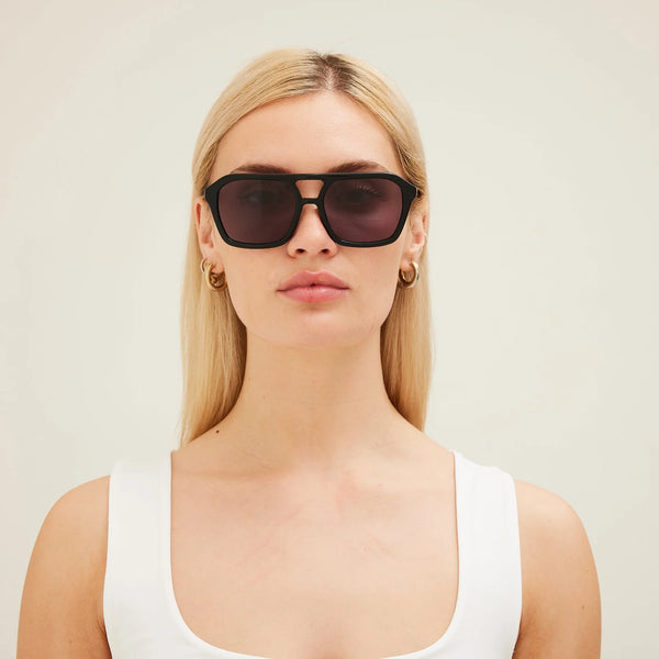 A front-on studio model shot featuring a blonde model with gold hoop earrings looking directly into the camera. She is wearing a plain white tank top and "The Void" sunglasses in a solid black frame colorway with dark gray lenses, highlighting the clean fit and symmetrical double bridge design.