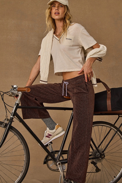 Side-angle lifestyle portrait of a model sitting on a bicycle, wearing the metallic chocolate wide-leg pant and a white t-shirt.