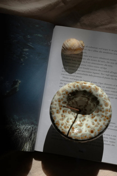Top-down lifestyle shot of the Gentle Habits Vessel Incense Holder in Seafoam resting on an open art book next to a small seashell.