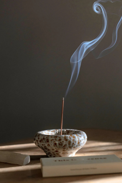 Mood shot of a lit incense stick burning in the Gentle Habits Vessel Incense Holder in Seafoam with smoke rising against a dark, warm-lit background.