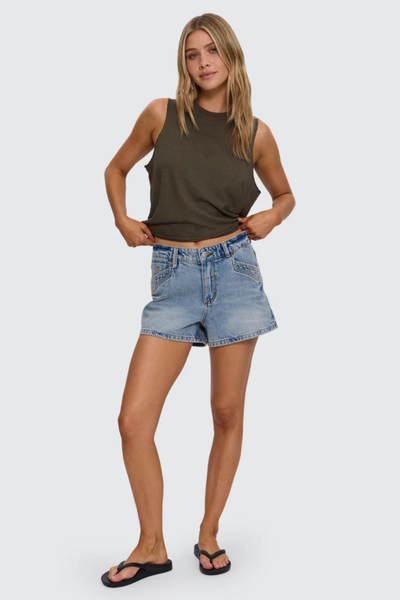 Studio full-length front portrait of a blonde model wearing sun-faded light blue mid-rise denim shorts with western-style front pockets, styled with an olive green tank top and black flip-flops.