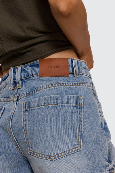 Close-up studio rear detail shot showing the brown leather Thrills logo waistband patch, double back belt loops, and back pocket contrast stitching on light blue denim shorts.