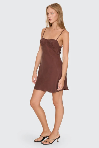 Side three-quarter view of a blonde model wearing the Thrills Elio Bias Mini Dress in Cocoa, showcasing the mini length, spaghetti straps, and gathered bust detail paired with black strappy heels.