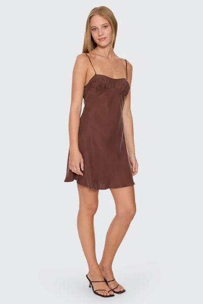 Full-length front view of model posing in the brown Thrills Elio Bias Mini Dress in Cocoa with slim shoulder straps and black strappy low heel mules against a plain white background.