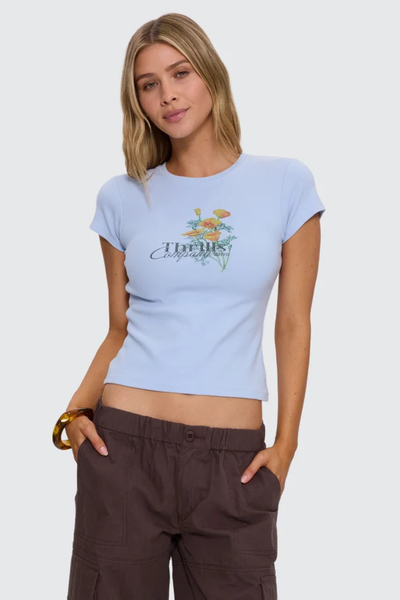 Mid-shot studio front portrait of a model wearing a fitted placid light blue fine rib organic cotton tee with a floral chest graphic, styled with brown pants.