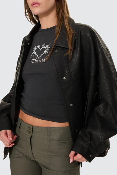 An outdoor winter lifestyle image showing a model from the front wearing the open black Stevie Jacket layered over a simple white top, styled casually with dark utility trousers against a blurred street backdrop.