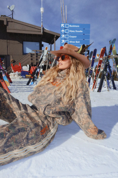 An outdoor lifestyle campaign image capturing a model from the side in a snowy mountain setting, wearing the oversized tree camo hoodie styled with black winter pants and a beanie, emphasizing its cold-weather utility and relaxed fit.