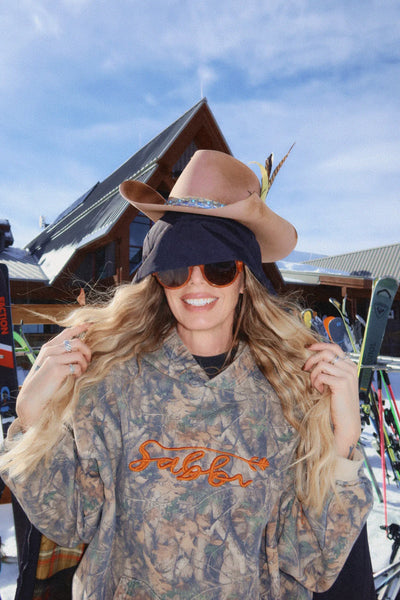 A candid outdoor lifestyle shot of a model sitting on a ski resort bench wearing the Sabbi tree camo fleece hoodie, showcasing the casual, streetwear-inspired drape of the 100% cotton fabrication in a real-world winter environment.