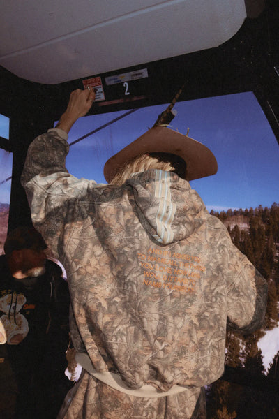 A candid outdoor lifestyle shot of a model sitting on a ski resort bench wearing the Sabbi tree camo fleece hoodie, showcasing the casual, streetwear-inspired drape of the 100% cotton fabrication in a real-world winter environment.