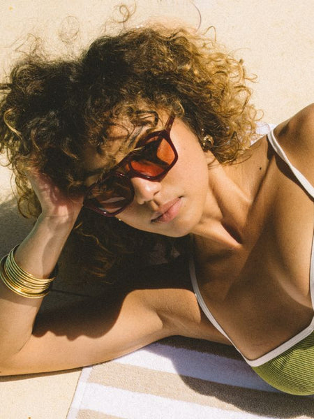 A sun-drenched outdoor campaign photograph of a model with curly hair sunbathing poolside on a striped beach towel. Looking down at the camera while leaning on her arm, she wears the translucent berry aviator sunglasses with a green swim top and gold bangle bracelets, capturing a relaxed summer lifestyle feel.