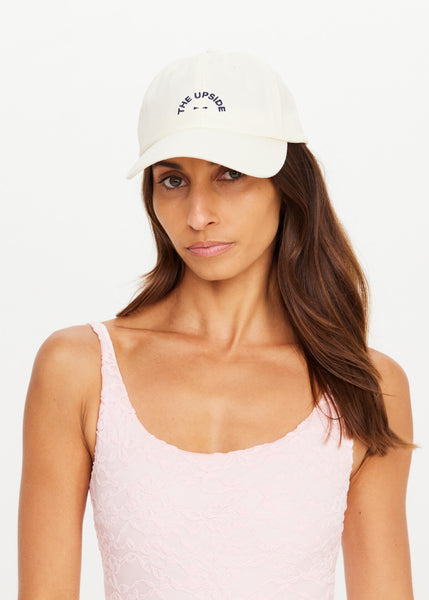 Front portrait studio shot of a model wearing The Upside Marlon Soft Cap in cream. The classic-fit cotton cap features a curved brim and navy blue embroidered arched branding on the front panel, styled with a light pink lace athletic bodysuit.