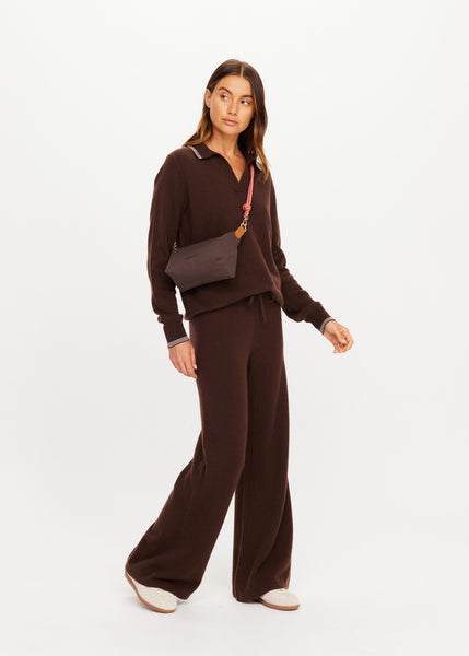 Full-length standing view of model Eden wearing a brown knit lounge set and wearing The Upside Poppy Crossbody Bag in Chocolate with a coral pink paracord strap across her chest.