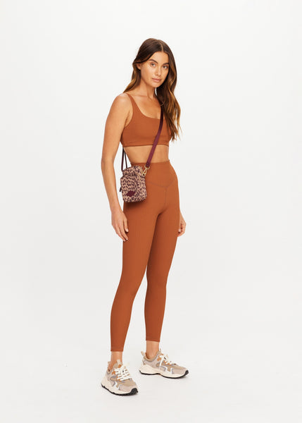 Full-length studio shot of a model styling The Upside Javan Lilo Crossbody Bag in leopard print. The compact, recycled polyester bag is worn across the body with its adjustable burgundy strap, accessorized alongside a matching ribbed terracotta activewear set and neutral-toned athletic sneakers.