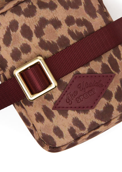 A detailed, close-up product shot of The Upside Javan Lilo Crossbody Bag in leopard print. The image highlights the rich texture of the recycled fabric, the gold-toned metal adjustment buckle, the ribbed burgundy webbing strap, and the stitched faux-leather diamond patch featuring embossed "The Upside SPORT" branding.
