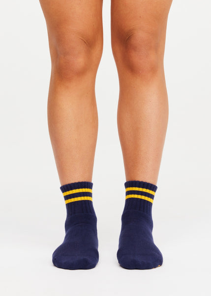 Front studio view of a model’s feet and lower legs wearing The Upside Salvador Grip Socks. The image highlights the classic navy cotton-blend knit, comfortable ribbed ankle cuffs detailed with two sporty lemon yellow stripes, and a supportive, snug fit ideal for Pilates, yoga, or barre workouts.