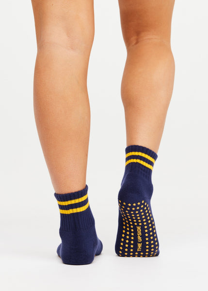 Rear studio view of a model’s lower legs wearing The Upside Salvador Grip Socks in navy blue. The shot showcases the midi ankle height with two prominent lemon yellow horizontal stripes on the ribbed cuff, and highlights the anti-slip sole covered in star-shaped yellow grippers alongside the printed brand name "THE UPSIDE".