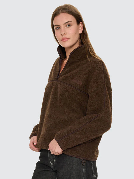 A mid-shot photograph focusing on the front design of the brown sherpa fleece pullover, detailing the premium brand logo embroidery on the left chest, functional zipped hand pockets, and the clean finish of the contrasting black hardware.