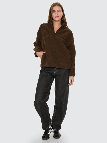 An outdoor winter lifestyle image featuring a model from the front wearing the brown All Star Sherpa Fleece, styled with dark vintage denim jeans against a soft-focus urban street backdrop.