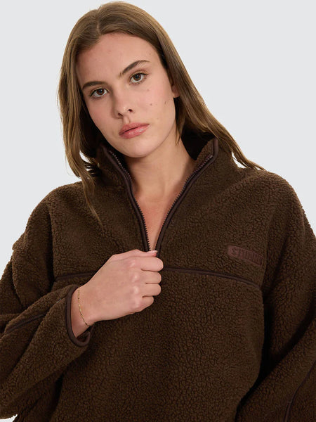 A candid mid-length outdoor photograph capturing the model looking away, highlighting the deep chocolate brown tone of the sherpa fleece jumper and its utility-inspired zipped pocket layout in a real-world environment.