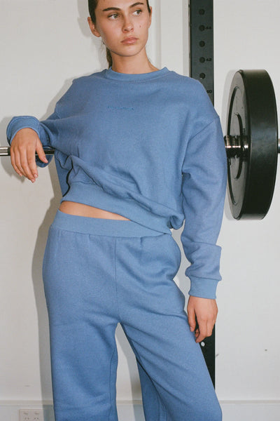 A campaign image featuring a model posing next to a barbell weight rack, wearing the coordinating blue organic cotton and hemp fleece crewneck sweatshirt and relaxed-fit wide sweatpants.