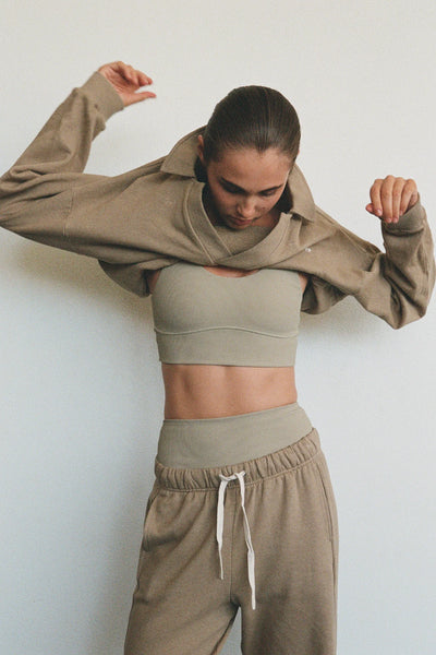 A candid campaign snapshot showing a model adjusting her hair while pulling on the olive collared sport sweatshirt, emphasizing the flexible stretch and soft texture of the premium mid-weight French terry fabric.