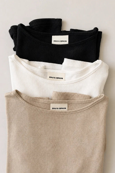 Flat lay view of three stacked Zulu & Zephyr Rib Hemp Jersey tops in beige husk, white, and black, displaying the bound crew necklines and inner woven brand labels.